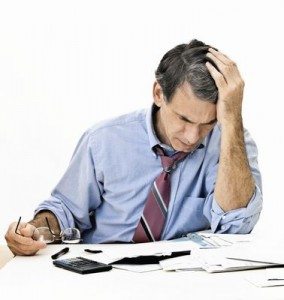 Man at desk in shirt and tie holding his head and worrying about money.